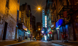 In Chinatown Rome, the streets are quiet at night with illuminated building signs on one side of the road giving the impression that they are open, while businesses on the other side appear closed without any lights.