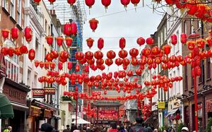 Red lanterns hang above a bustling crowd of people in London's Chinatown, with individuals occupying both sides of the gathering.